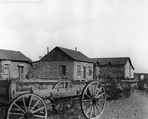 A view of 3 adobe buildings with peaked rooves with at flat bed wagon in the foreground.